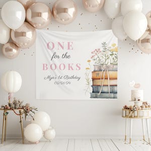 May include: A white banner with the text "ONE for the BOOKS" in pink lettering. The banner also includes the text "Mya's 1st Birthday 02/21/24" and a watercolor illustration of a stack of books with flowers.