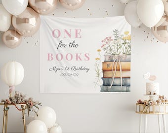 May include: A white banner with the text "ONE for the BOOKS" in pink lettering. The banner also includes the text "Mya's 1st Birthday 02/21/24" and a watercolor illustration of a stack of books with flowers.