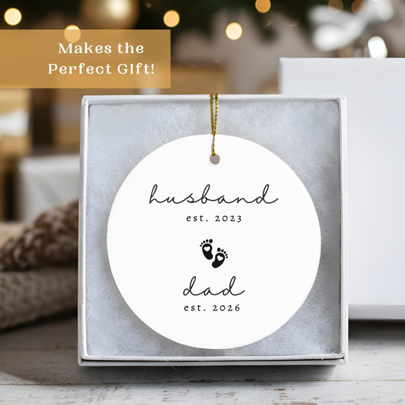 May include: A white, round ceramic ornament with the words "husband est. 2023" and "dad est. 2026" in black script, along with baby footprints. The ornament is in a white gift box with a gold string for hanging.