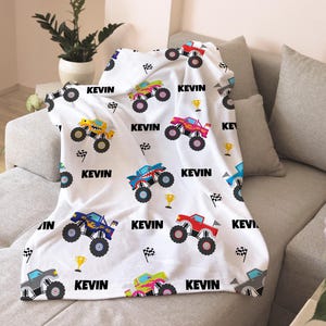 May include: White fleece blanket with a repeating pattern of monster trucks in various colors. The trucks have names and numbers on them. The blanket has the name "Kevin" printed on it.