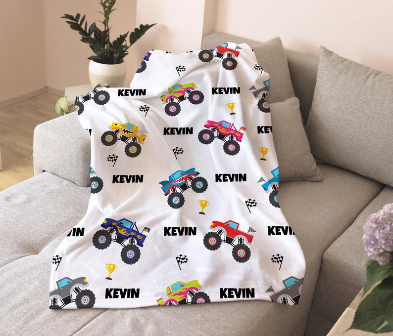 May include: White fleece blanket with a repeating pattern of monster trucks in various colors. The trucks have names and numbers on them. The blanket has the name "Kevin" printed on it.