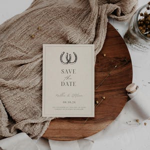 K&ouml;nnte beinhalten: Eine rustikale Save-the-Date-Karte mit braunem Hintergrund und schwarzem Text. Die Karte zeigt zwei Hufeisen und den Text "Save the Date" mit den Namen "Kalie & Steffen" und dem Datum "08.10.26".