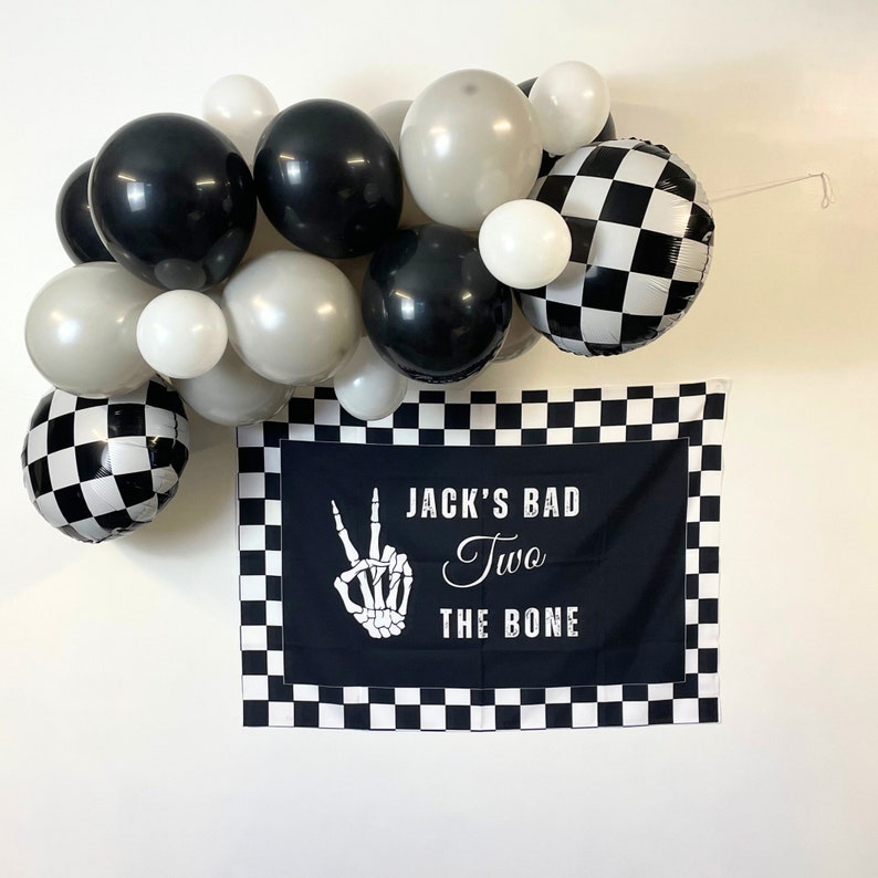 May include: Black and white checkered balloons and a banner with the text "Jack's Bad Two The Bone" and a skeleton hand making a peace sign.