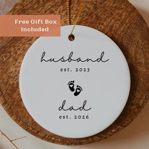May include: A white ceramic ornament with the words "husband est. 2023" and "dad est. 2026" in black script. The ornament features black baby footprints. A gold string is attached for hanging. The words "Free Gift Box Included" are in a box in the upper left corner.
