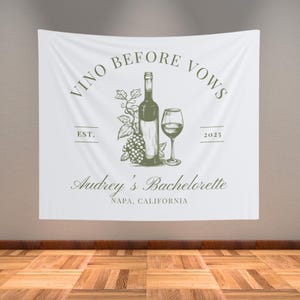 May include: White banner with the text "Vino Before Vows" in green lettering. The banner also features a drawing of a bottle of wine, a glass of wine, and grapes. The text "Audrey's Bachelorette" is below the drawing, along with the location "Napa, California".