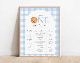 May include: A framed baby's first birthday milestone poster with a light blue gingham background. The poster includes the text "ASHER'S ONE sweet year" and details such as height (29 inches), weight (21 pounds), and favorite foods.