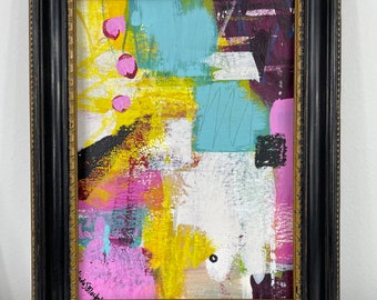 May include: Abstract painting in a black frame with a gold trim. The painting features a mix of bright colors, including yellow, pink, blue, and white. The artist's signature is visible in the bottom left corner.