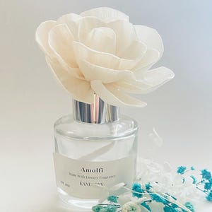 May include: A clear glass fragrance diffuser bottle with a silver collar and a large, cream-coloured sola wood flower topper. The label reads Amalfi, Made With Luxury Fragrance, KANEDY, EST 2021. Small white and teal dried flowers sit at the base.