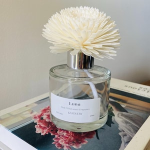 May include: A clear glass diffuser bottle with a silver cap and a large, cream-coloured flower-shaped diffuser. The bottle has a white label with the word "Luna" and "KANDLERY" printed on it. The diffuser sits on a book with pink flowers.