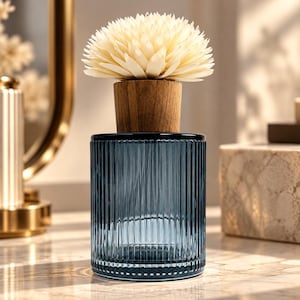May include: A cylindrical blue glass diffuser bottle with vertical ribbed texture, topped with a light brown wooden stopper and a cream-coloured, multi-petaled flower-shaped reed diffuser attachment made of thin, layered material.