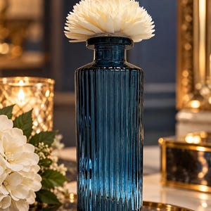 May include: A tall, cylindrical, dark blue glass bottle with vertical ribbed texture throughout its body. The bottle features a narrow neck and a flat rim, topped with a large, cream-coloured, multi-petaled decorative flower diffuser.
