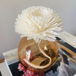 May include: A decorative diffuser featuring a large, cream-coloured flower atop a round, amber-coloured glass vessel. A white wick extends from the flower into the vessel. The diffuser rests on a stack of books.