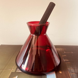 May include: A translucent ruby red glass vessel with a wide, conical base and a narrow, flared neck. A single dark brown, slender wooden reed stick is inserted into the opening of the glass container, extending diagonally upwards from the centre.