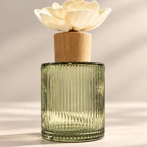 May include: A cylindrical olive green glass diffuser bottle with vertical ribbed texture, topped with a light-coloured wooden stopper and a large, cream-coloured decorative flower made of thin, layered petals extending from the centre.