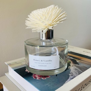 May include: A clear glass diffuser bottle with a silver neck and a large, cream-coloured flower-shaped diffuser. The label reads "Honey & Vanilla" and "KANDLERY". The bottle sits on a stack of books.