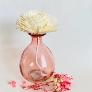 May include: A pink glass diffuser bottle with a white wooden flower diffuser on top. The bottle has a round shape and a clear glass bottom.