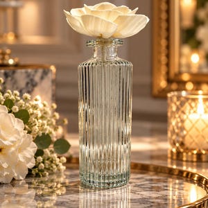 May include: A clear glass reed diffuser bottle with a vertical ribbed texture and a narrow neck. A large, cream-coloured, multi-petaled sola wood flower is inserted into the top of the bottle, serving as a decorative diffuser element.