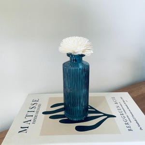 May include: A teal blue glass vase with vertical ribbed detailing, topped with a white, flower-shaped diffuser. The vase sits on a book with the text "MATISSE PAPIERS DÉCOUPÉS" and "BERGGRUEN & CIE".