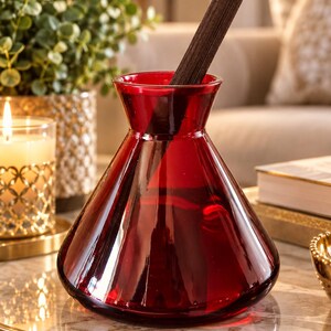 May include: A translucent, deep red glass vase with a wide, conical base that tapers upwards to a narrow, circular opening. A single, dark brown, textured wooden reed or stick is inserted into the top of the vase, extending outwards.