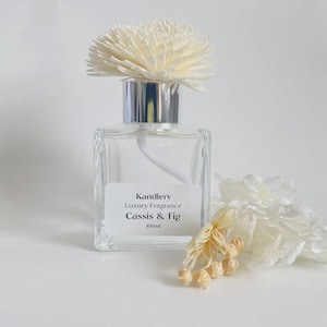 May include: A clear, square glass bottle with a silver cap and a white flower diffuser. The label reads "Kandlery Luxury Fragrance Cassis & Fig 100ml." A sprig of dried flowers rests beside the bottle, suggesting a floral fragrance.