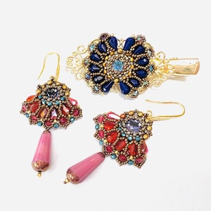 Può includere: Un set di gioielli composto da due orecchini pendenti e una molletta per capelli. Gli orecchini presentano cristalli rossi e rosa con una perla rosa a goccia. La molletta ha un design circolare con petali blu scuro e un cristallo centrale.