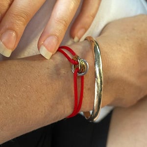 Może przedstawiać: Czerwona bransoletka ze sznurka ze srebrnym i złotym zapięciem w kształcie pierścienia. Widoczna jest również złota bransoletka. Bransoletki są na nadgarstku osoby.