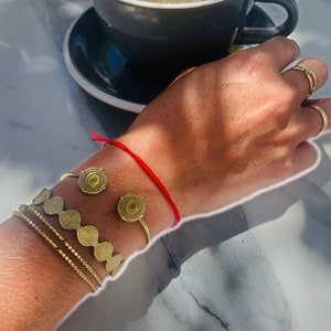 Può includere: Un braccio con un braccialetto di corda rossa e tre braccialetti d'oro con disegni circolari. I braccialetti sono impilati uno sopra l'altro.