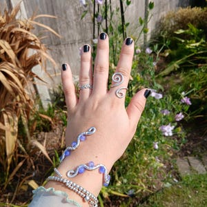 May include: A silver-toned wire wrap bracelet featuring small purple beads, a matching silver-toned spiral ring, a thin silver-toned band ring, and a multi-strand silver-toned beaded bracelet worn on the wrist and fingers.