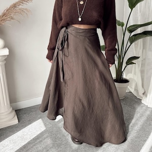 May include: A brown, cropped sweater and a wrap skirt. The sweater has a high neck and long sleeves. The skirt is a solid brown color and wraps around the waist with a tie. A layered necklace is visible.