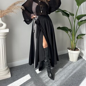May include: A black wrap skirt with a high slit and a black off-the-shoulder blouse. The blouse has long, flowing sleeves. The outfit is paired with black knee-high boots. The model is wearing sunglasses and layered necklaces.
