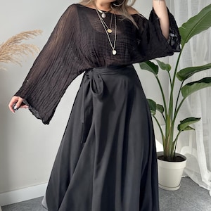 May include: A woman wearing a black off-the-shoulder top with long sleeves and a black wrap skirt. The outfit is accessorized with sunglasses and layered necklaces. The skirt is a midi length and the boots are black.