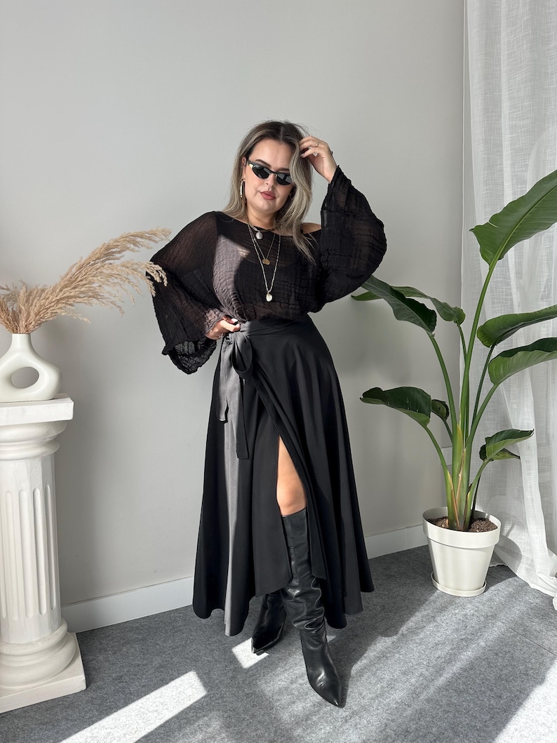 May include: A black wrap skirt with a high slit and a black off-the-shoulder blouse. The blouse has long, flowing sleeves. The outfit is paired with black knee-high boots. The model is wearing sunglasses and layered necklaces.