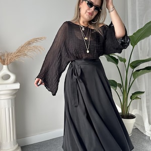 May include: A woman wearing a black outfit. The outfit includes a sheer, long-sleeved top and a long, flowing skirt with a wrap-around tie. She is wearing black sunglasses, jewelry, and black boots.