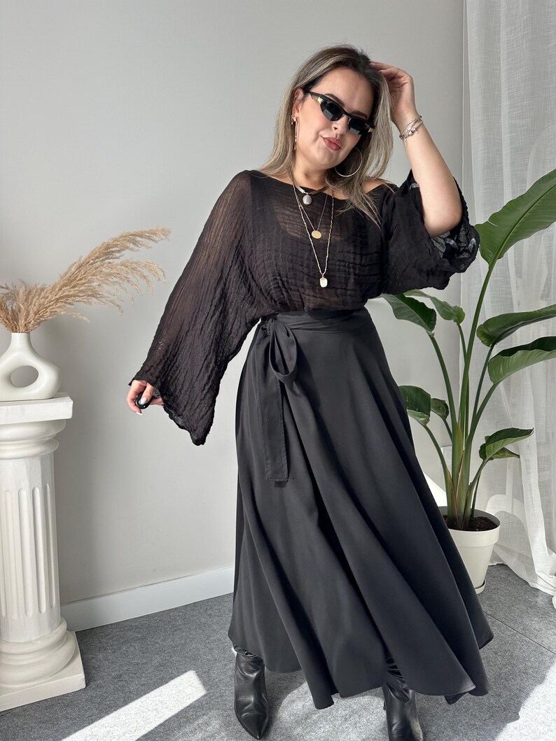 May include: A woman wearing a black outfit. The outfit includes a sheer, long-sleeved top and a long, flowing skirt with a wrap-around tie. She is wearing black sunglasses, jewelry, and black boots.