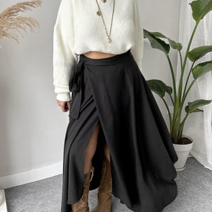 May include: A woman wearing a white, fuzzy sweater and a black wrap skirt. The skirt has a high slit and is paired with brown cowboy boots. The outfit is accessorized with layered necklaces and earrings.