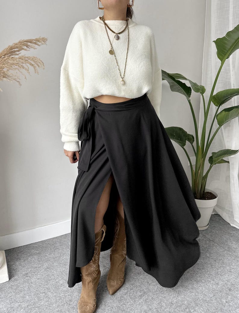 May include: A woman wearing a white, fuzzy sweater and a black wrap skirt. The skirt has a high slit and is paired with brown cowboy boots. The outfit is accessorized with layered necklaces and earrings.