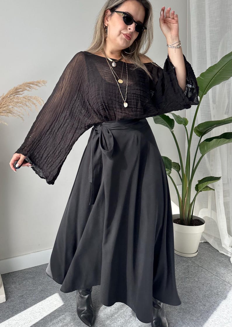 May include: A woman wearing a black off-the-shoulder top with long sleeves and a black wrap skirt. The outfit is accessorized with sunglasses and layered necklaces. The skirt is a midi length and the boots are black.