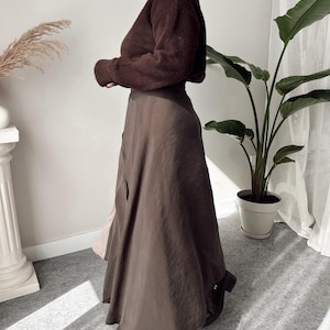May include: A woman wearing a brown sweater and a long, flowing brown skirt. The skirt has a subtle sheen and a slight A-line shape. The outfit is paired with black boots. The background includes a houseplant and a decorative column.
