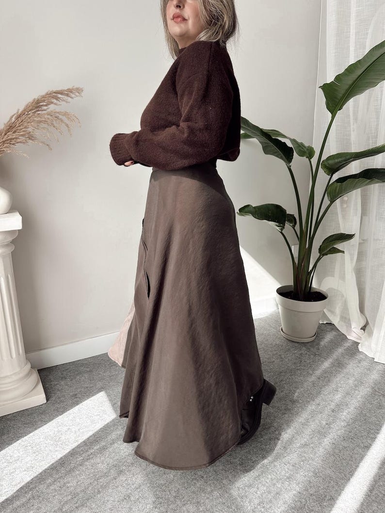 May include: A woman wearing a brown sweater and a long, flowing brown skirt. The skirt has a subtle sheen and a slight A-line shape. The outfit is paired with black boots. The background includes a houseplant and a decorative column.