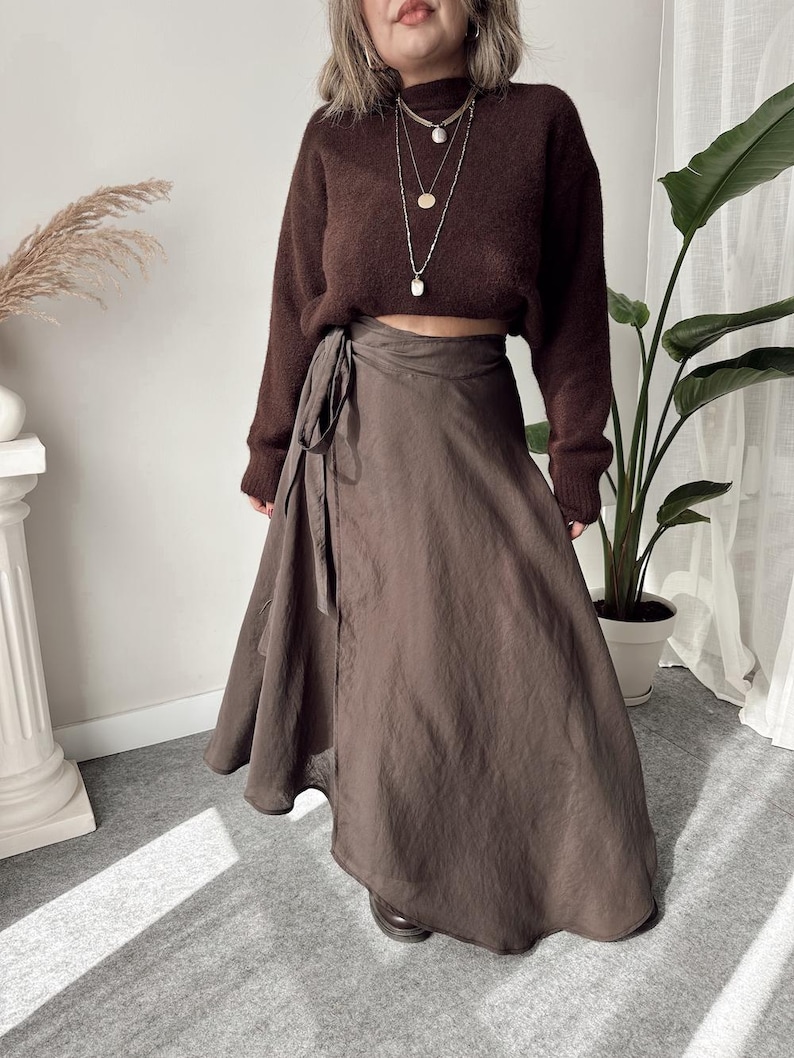 May include: A brown, cropped sweater and a wrap skirt. The sweater has a high neck and long sleeves. The skirt is a solid brown color and wraps around the waist with a tie. A layered necklace is visible.