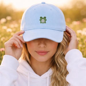 May include: Light blue baseball cap with a green frog embroidered on the front. The cap is made of a soft material and has a curved brim. The cap is being held by a person.
