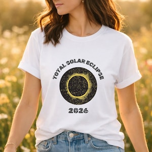 Puede incluir: Camiseta blanca con un gráfico negro y amarillo de un eclipse solar. El texto "TOTAL SOLAR ECLIPSE" está arqueado sobre el gráfico, con "2026" debajo. La camiseta está hecha de un material suave.