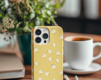 Puede incluir: Funda de teléfono amarilla con un estampado de patos blancos con sombreros azules. La funda del teléfono está sobre una mesa de madera junto a una taza de café, un libro y bolígrafos. El fondo incluye vegetación.