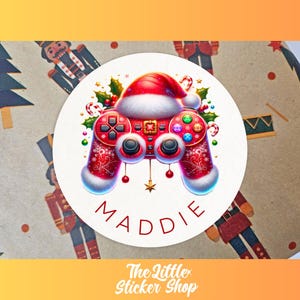 May include: A circular sticker with a festive design. It features a red and white video game controller adorned with a Santa hat and holly. The name "MADDIE" is displayed. The background has a pattern of nutcrackers and Christmas trees.