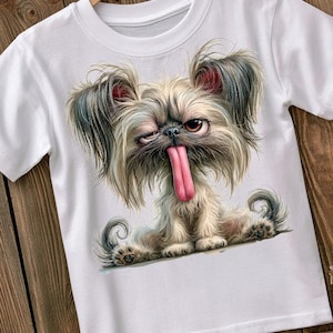 May include: A white short-sleeved t-shirt featuring a graphic illustration of a fluffy, long-haired dog with a scruffy appearance, one eye squinting, and a long pink tongue hanging out. The dog has grey and tan fur and is sitting down.