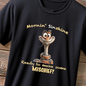 May include: A black short-sleeved t-shirt featuring a cartoon snake with wide eyes and a smirk. Above the snake is the text "Mornin' Sinshine" and below it is the text "Ready to make some MISCHIEF?" in a light yellow, sans-serif font.