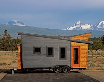 May include: A small, modern, gray and wood-paneled house on wheels with a bright orange door. The house is parked in a field with snow-capped mountains in the background.