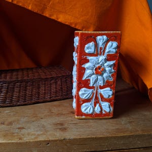 May include: Orange and white ceramic vase with a floral design. The vase is rectangular in shape and features white flowers and leaves on an orange background.