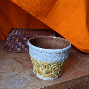 May include: A vintage ceramic planter in cream and brown. It has a scalloped rim and an embossed floral pattern. A woven basket is in the background, set against an orange fabric.