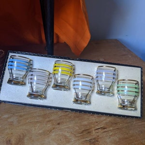 May include: Set of six clear glass shot glasses with gold rims and coloured stripes. The glasses are arranged in a black box with a white interior.